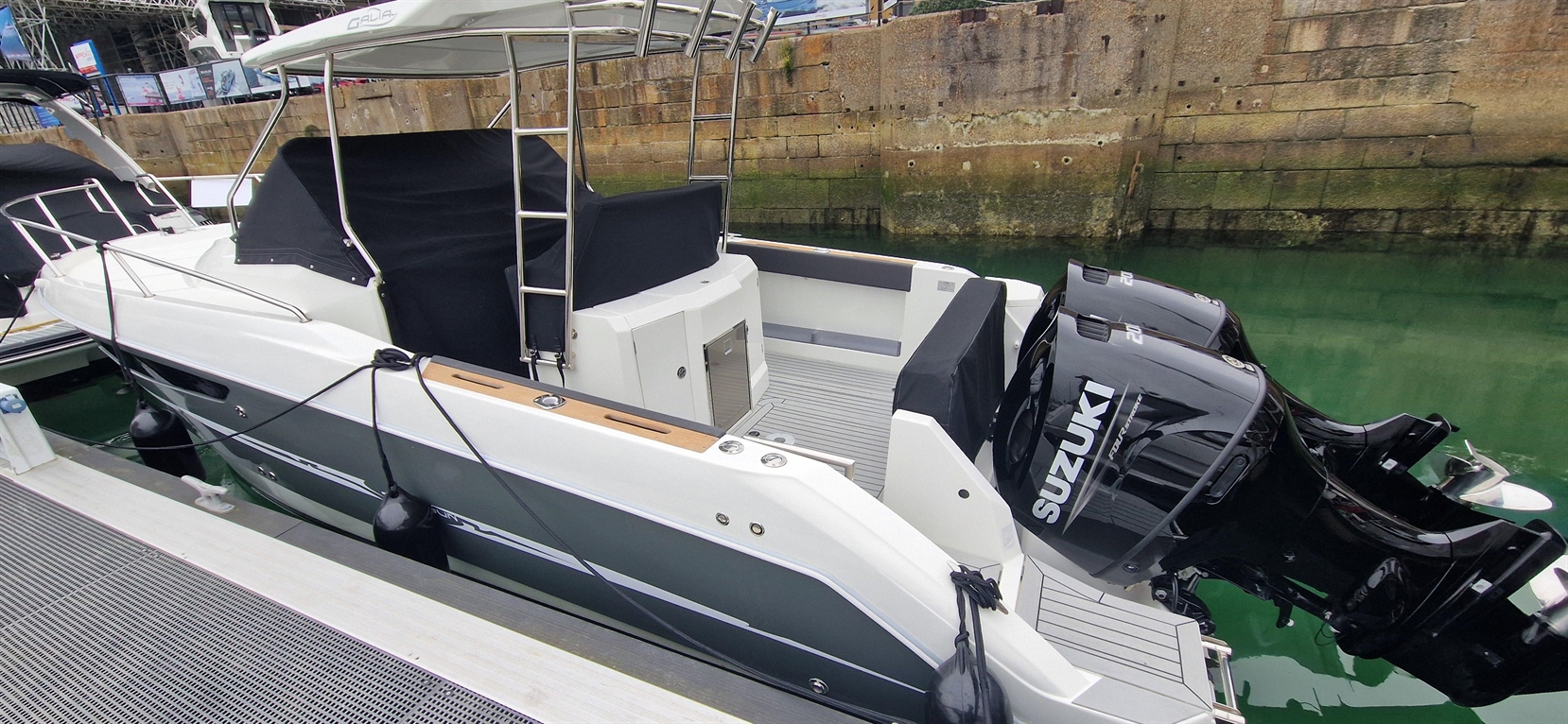 A sleek motorboat docked near a stone wall, featuring two Suzuki outboard motors and a spacious deck with gray flooring.